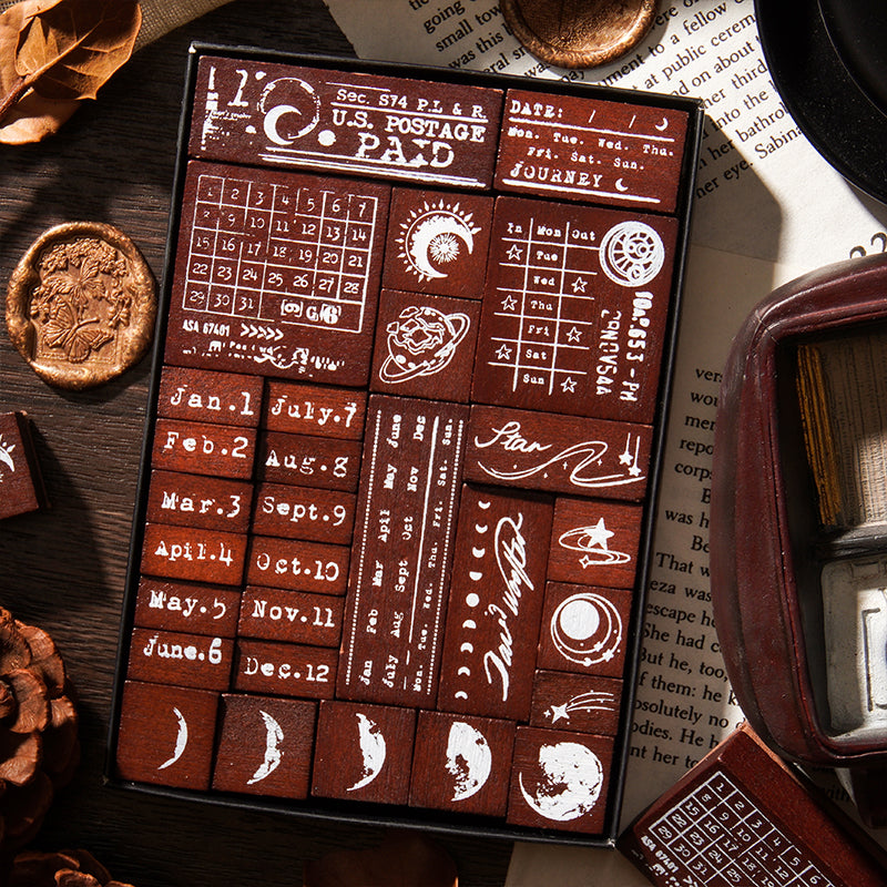 Close-up view of the 29 solid wood stamps packed neatly in the display box. Features highly detailed designs like vintage U.S. Postage Paid marks, retro calendars, and celestial moon phases for junk journaling.