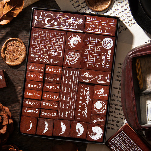Close-up view of the 29 solid wood stamps packed neatly in the display box. Features highly detailed designs like vintage U.S. Postage Paid marks, retro calendars, and celestial moon phases for junk journaling.