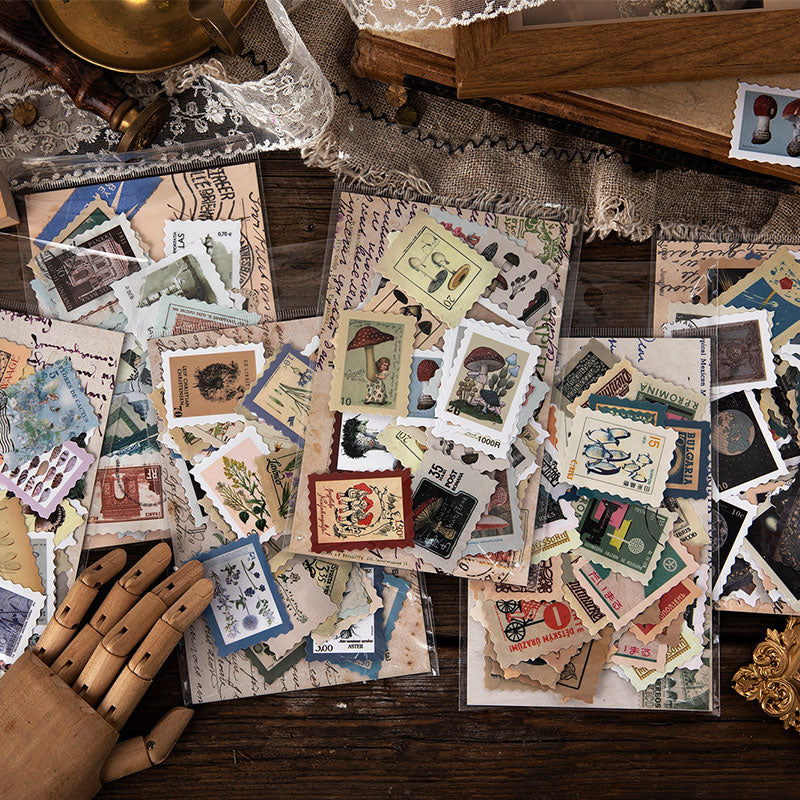 Six vintage postage stamp sticker packs scattered on dark wood desk with lace, showing botanical space and travel themes.