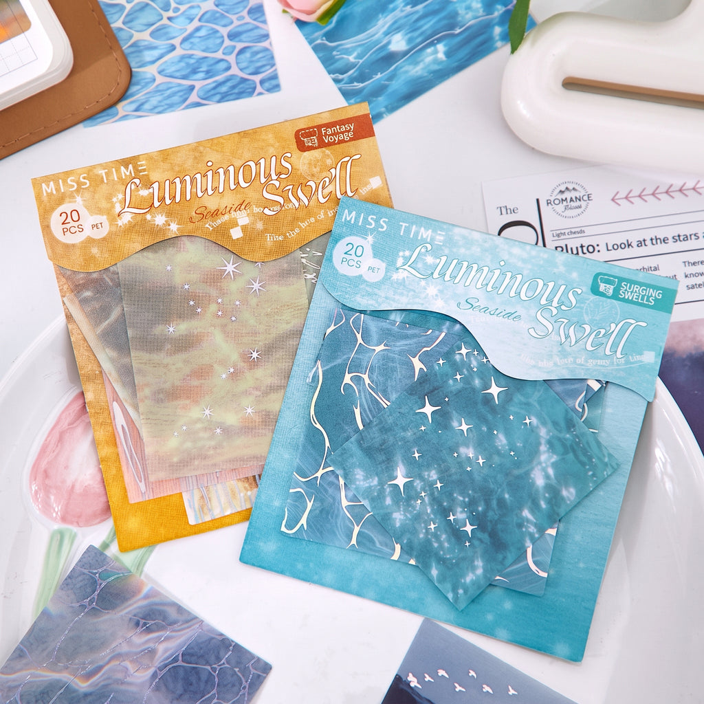 Two Luminous Swell water ripple sticker packs in gold and teal on white desk with journal supplies