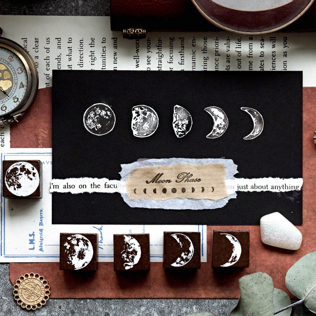 Close-up of white ink moon phase stamps beautifully layered on a black card and torn vintage paper. Five solid wood moon phase stamps are lined up below, showcasing creative mystic scrapbooking techniques.