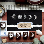 Close-up of white ink moon phase stamps beautifully layered on a black card and torn vintage paper. Five solid wood moon phase stamps are lined up below, showcasing creative mystic scrapbooking techniques.