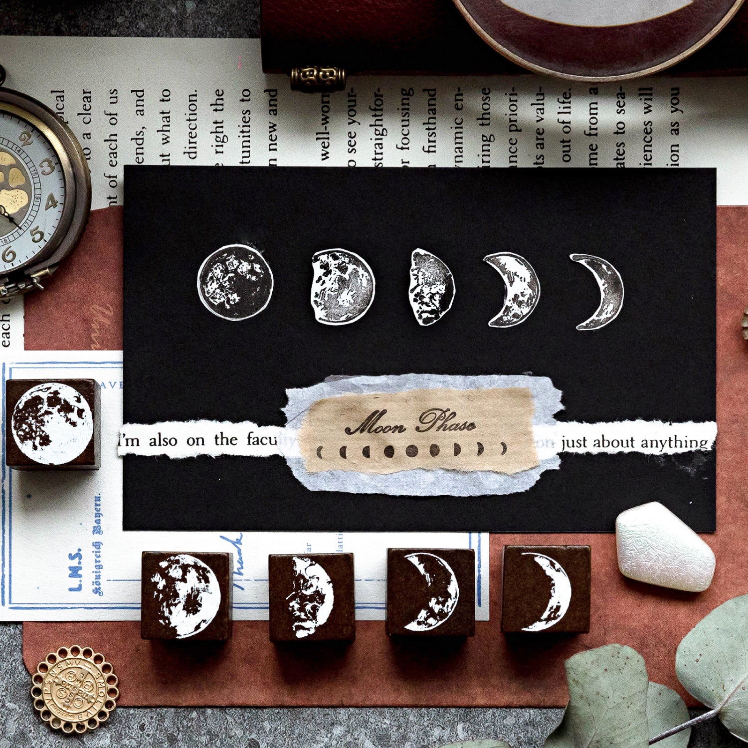 Close-up of white ink moon phase stamps beautifully layered on a black card and torn vintage paper. Five solid wood moon phase stamps are lined up below, showcasing creative mystic scrapbooking techniques.